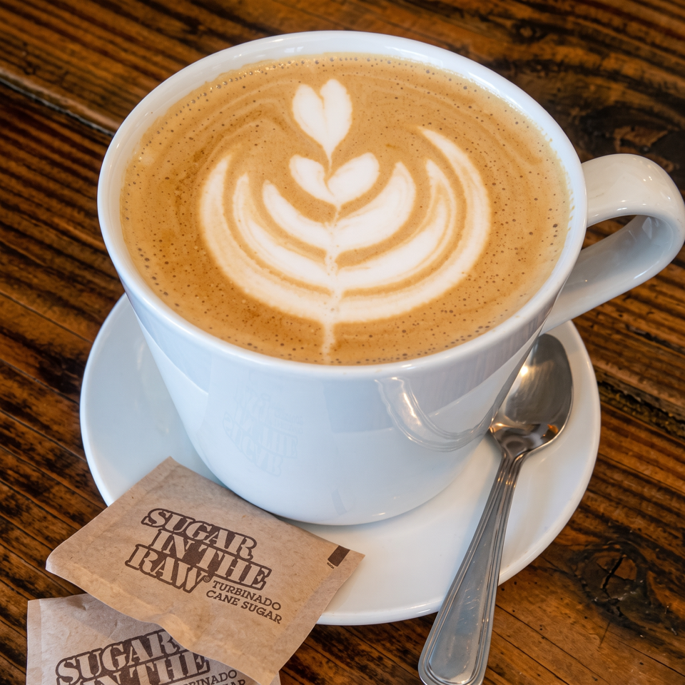 Latte with leaf-shaped foam art in a white cup, saucer, and spoon on a wooden table beside Sugar in the Raw packets.